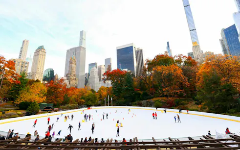People ice skating at Wollman Rink in Central Park, NYC, with autumn foliage and skyscrapers in the background.