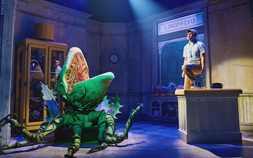 Man standing on counter next to large plant puppet in "Little Shop of Horrors" stage set.