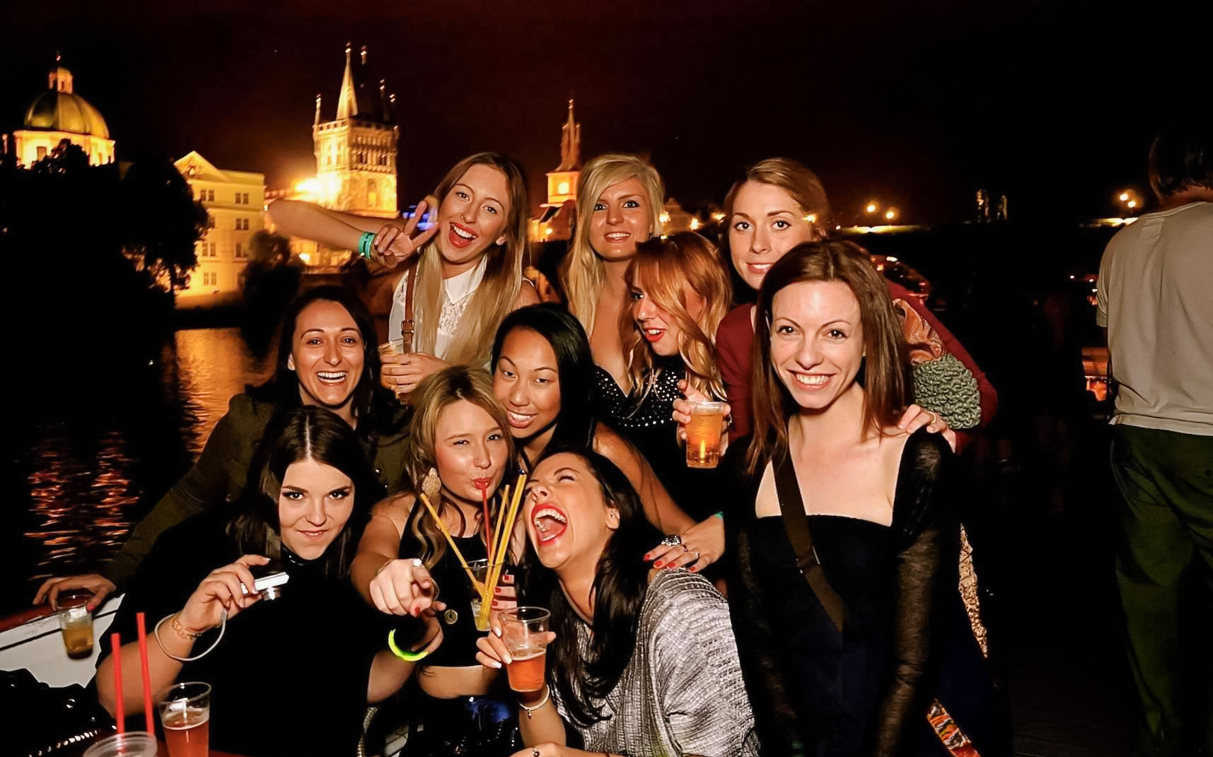 Group enjoying a boat party at night with Prague landmarks in the background.