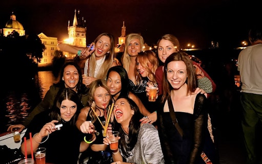 Group enjoying a boat party at night with Prague landmarks in the background.