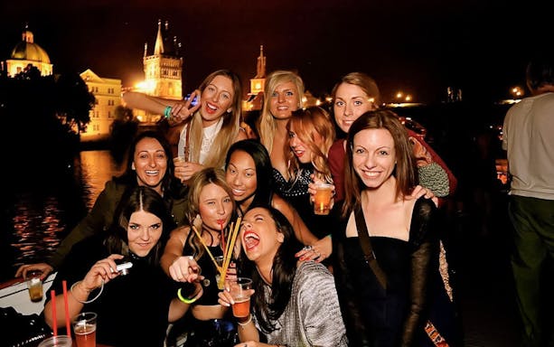 Group enjoying a boat party at night with Prague landmarks in the background.
