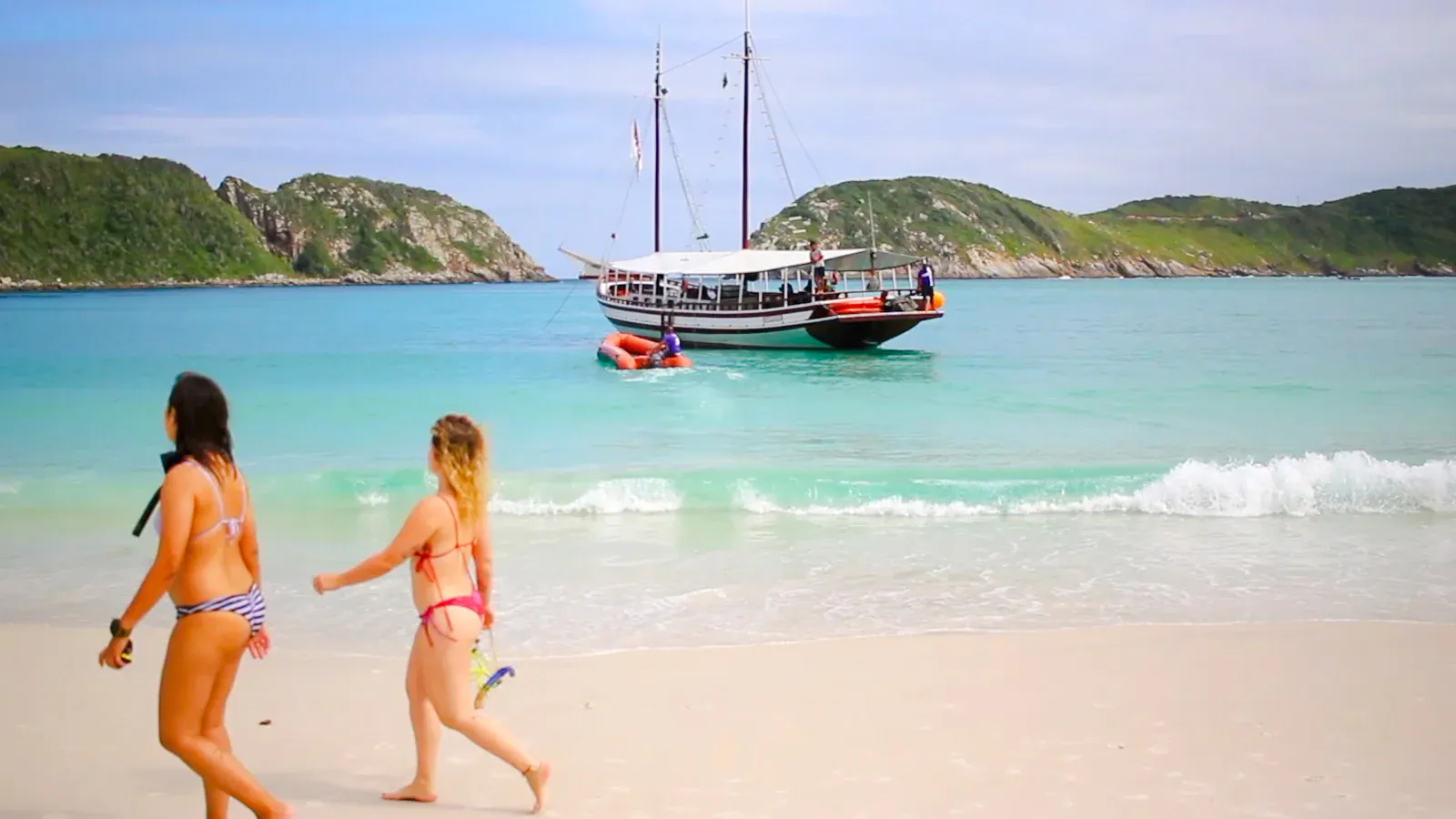Tourists walking on a beach with a boat in the turquoise waters of Arraial do Cabo, Brazil.