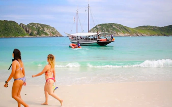 Tourists walking on a beach with a boat in the turquoise waters of Arraial do Cabo, Brazil.