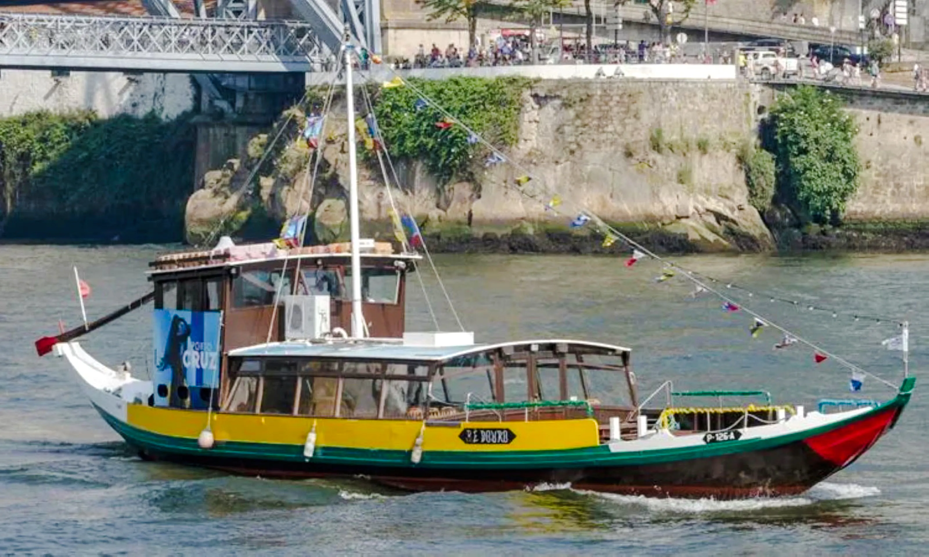 Traditional boat cruising on the Douro River in Porto, near a stone embankment and bridge.