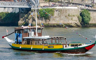 Traditional boat cruising on the Douro River in Porto, near a stone embankment and bridge.