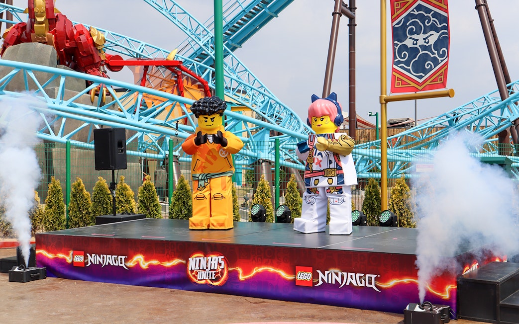 Lego Ninjago characters on stage at Legoland with roller coaster in background.
