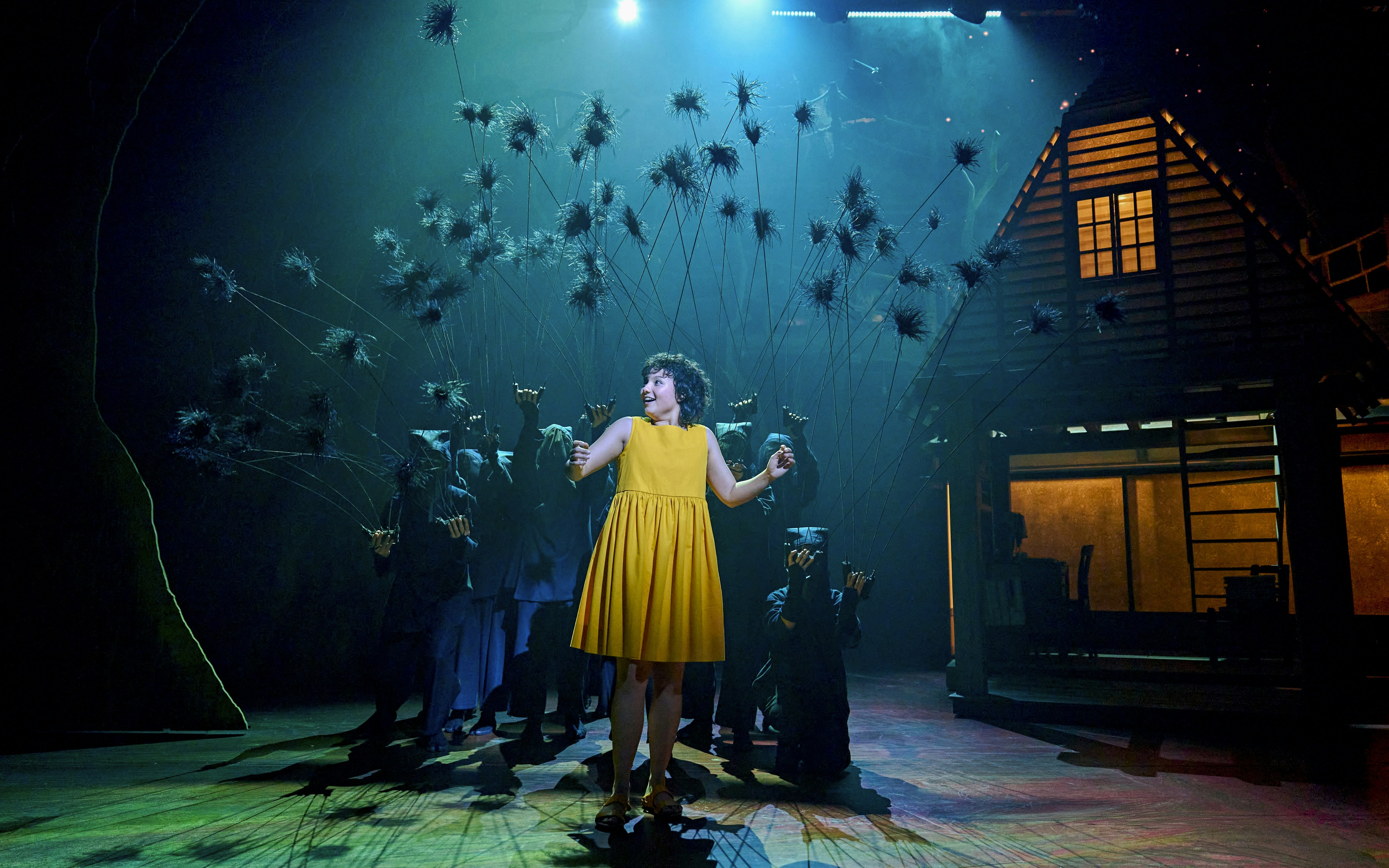 Stage scene from My Neighbour Totoro with a performer in a yellow dress surrounded by puppeteers.