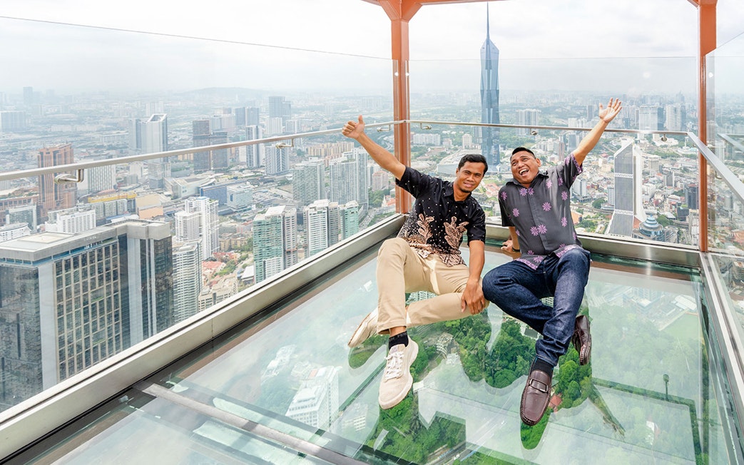 Tourists sitting on the Sky Glass box at Kuala Lumpur Tower with city skyline view.