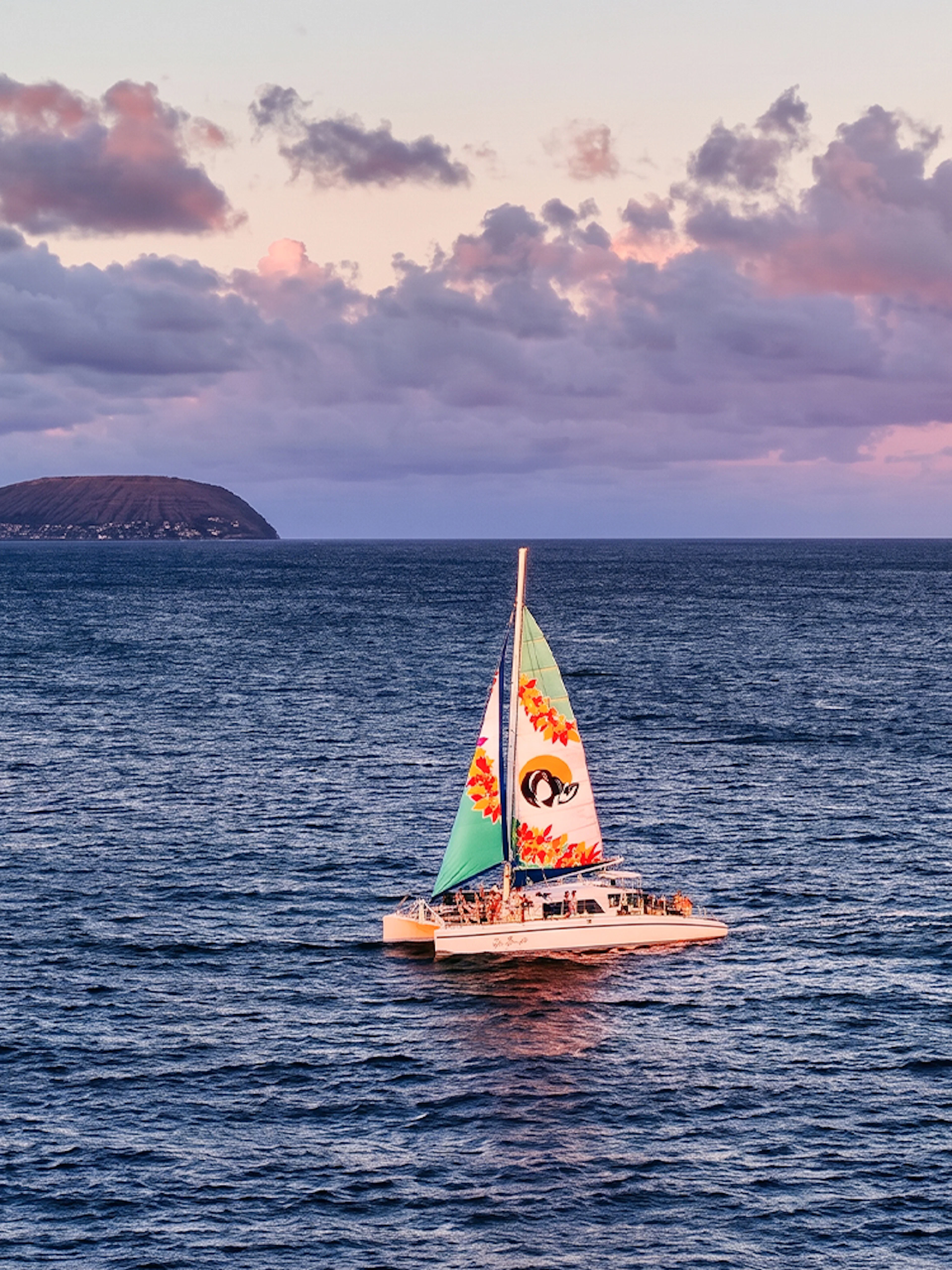 Catamaran sailing at sunset near Oahu, Hawaii, with distant islands in view.