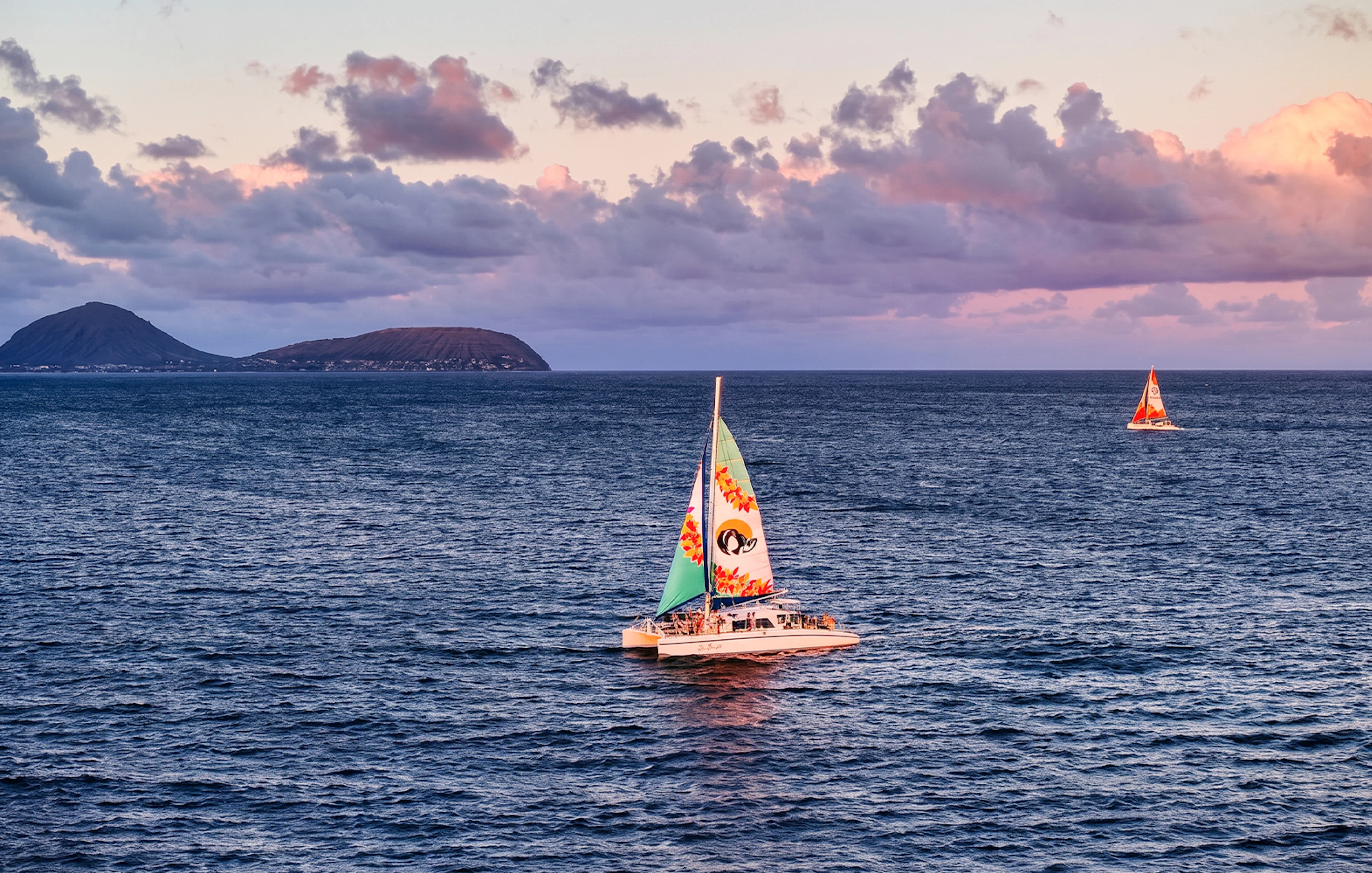 Catamaran sailing at sunset near Oahu, Hawaii, with distant islands in view.