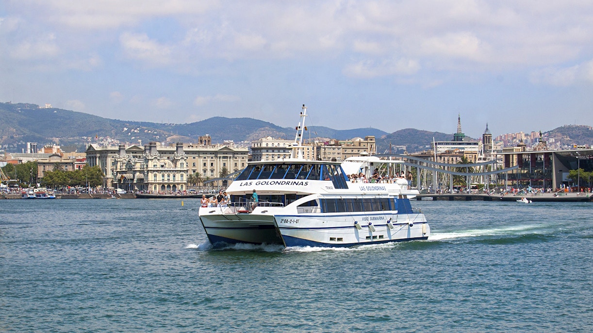 Las Golondrinas boat cruising Barcelona coastline with city skyline in background.