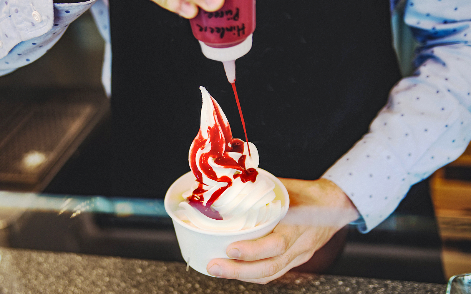 Seller pouring red sauce on soft frozen yogurt in a white takeaway cup.