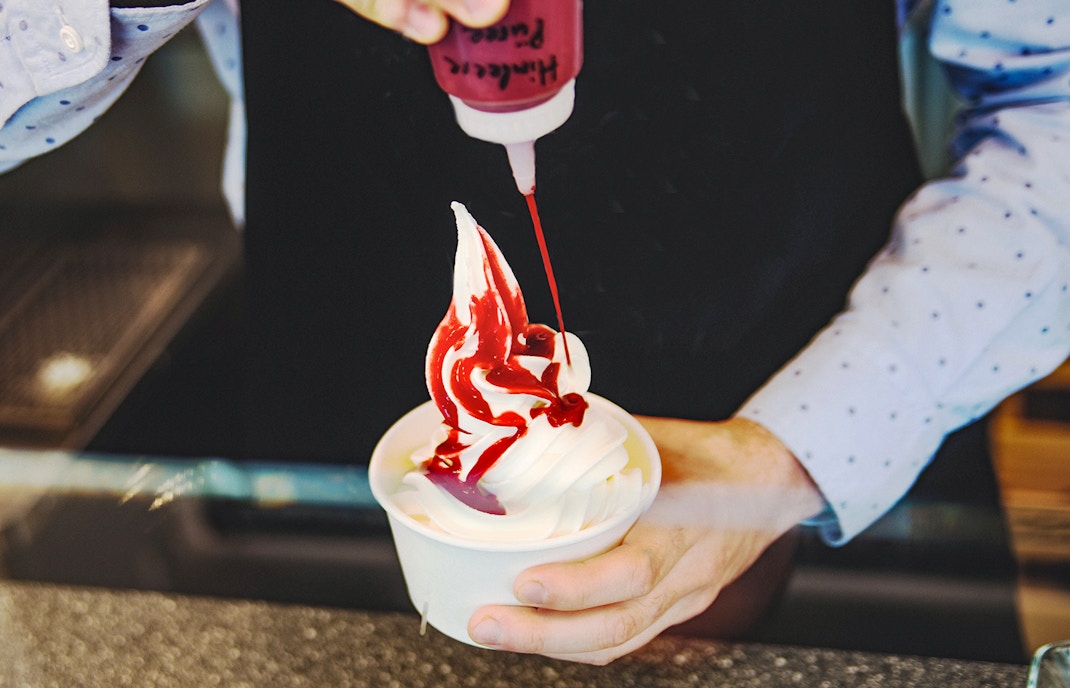 Seller pouring red sauce on soft frozen yogurt in a white takeaway cup.