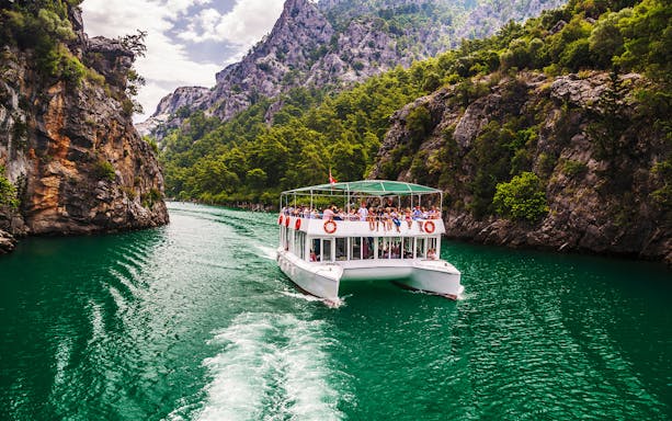 Boat navigating through Green Canyon with tourists, surrounded by rocky cliffs and lush greenery in Antalya.