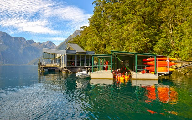 Milford Sound dock with kayaks and observatory, surrounded by mountains and forest.