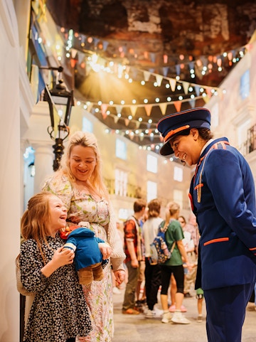 Child holding Paddington Bear toy, smiling with adult and costumed guide at Paddington Bear Experience.