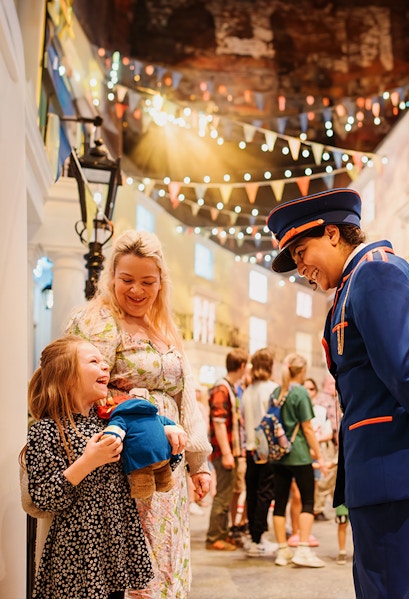 Child holding Paddington Bear toy, smiling with adult and costumed guide at Paddington Bear Experience.