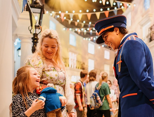 Child holding Paddington Bear toy, smiling with adult and costumed guide at Paddington Bear Experience.