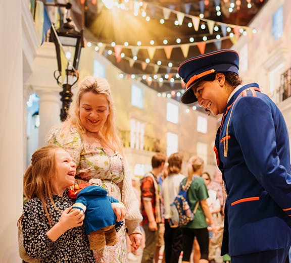 Child holding Paddington Bear toy, smiling with adult and costumed guide at Paddington Bear Experience.
