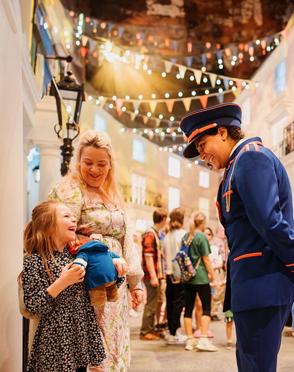 Child holding Paddington Bear toy, smiling with adult and costumed guide at Paddington Bear Experience.
