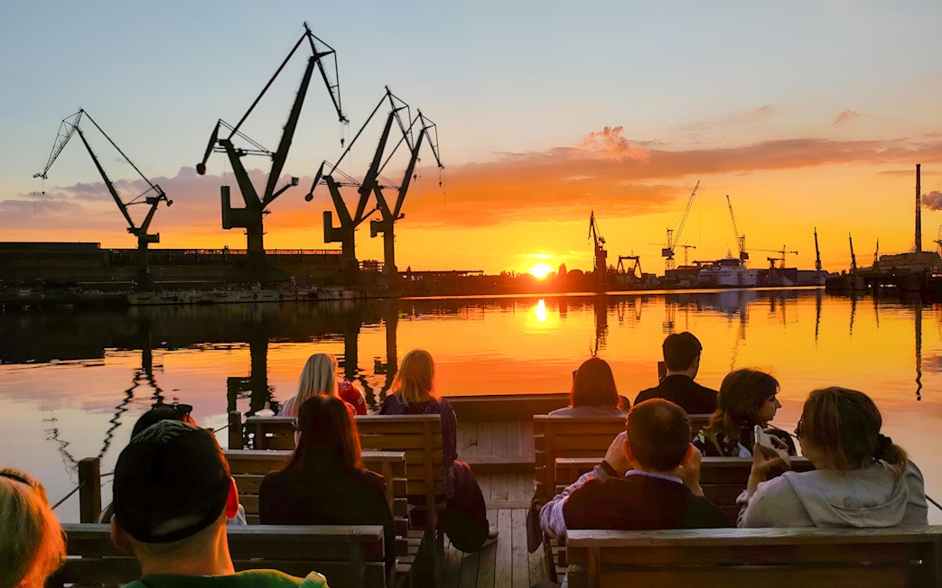 People on a river cruise in Gdańsk enjoying sunset with shipyard cranes in view.