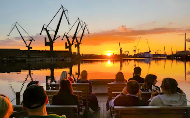 People on a river cruise in Gdańsk enjoying sunset with shipyard cranes in view.