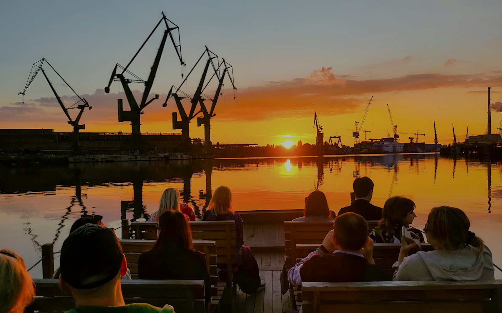 People on a river cruise in Gdańsk enjoying sunset with shipyard cranes in view.