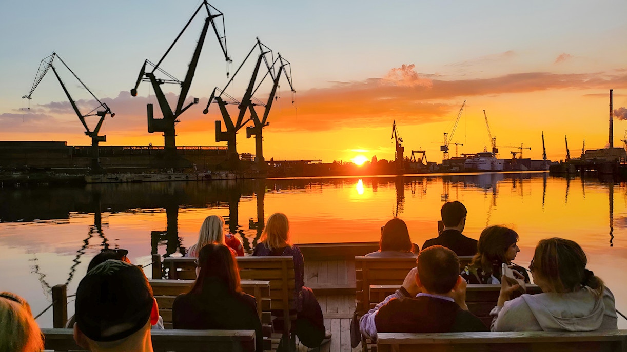 People on a river cruise in Gdańsk enjoying sunset with shipyard cranes in view.