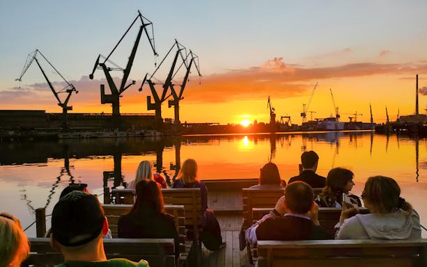 People on a river cruise in Gdańsk enjoying sunset with shipyard cranes in view.