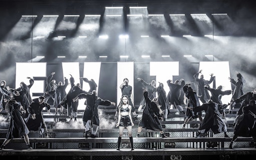 Performers on stage during Evita musical with large illuminated letters in the background.