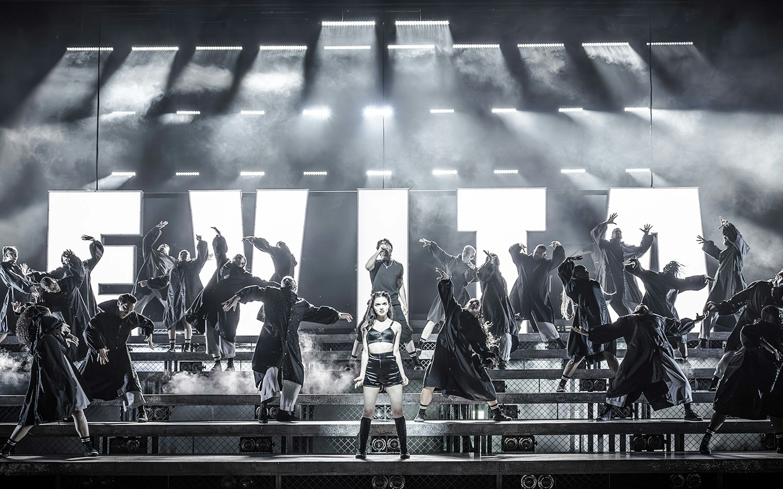 Performers on stage during Evita musical with large illuminated letters in the background.