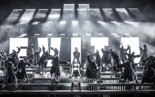 Performers on stage during Evita musical with large illuminated letters in the background.