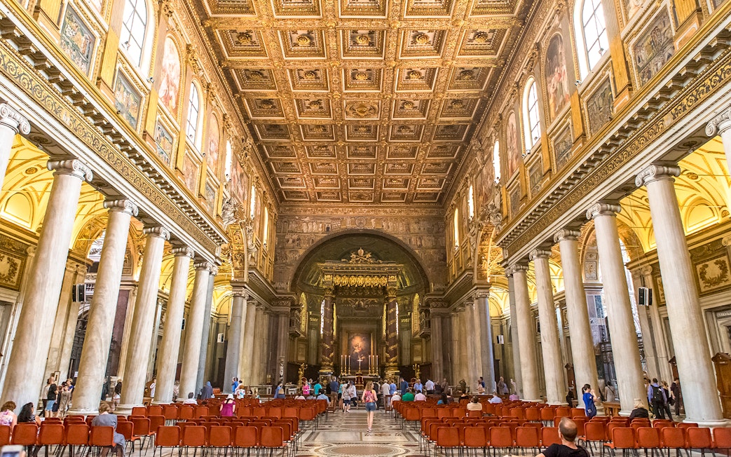 Visitors at the altar of Basilica Santa Maria Maggiore, Rome, with ornate ceiling and columns.