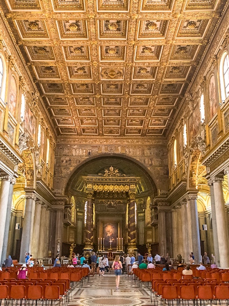 Visitors at the altar of Basilica Santa Maria Maggiore, Rome, with ornate ceiling and columns.