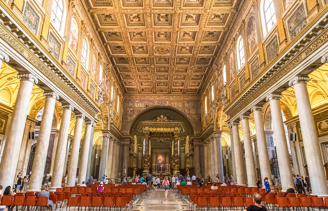 Visitors at the altar of Basilica Santa Maria Maggiore, Rome, with ornate ceiling and columns.