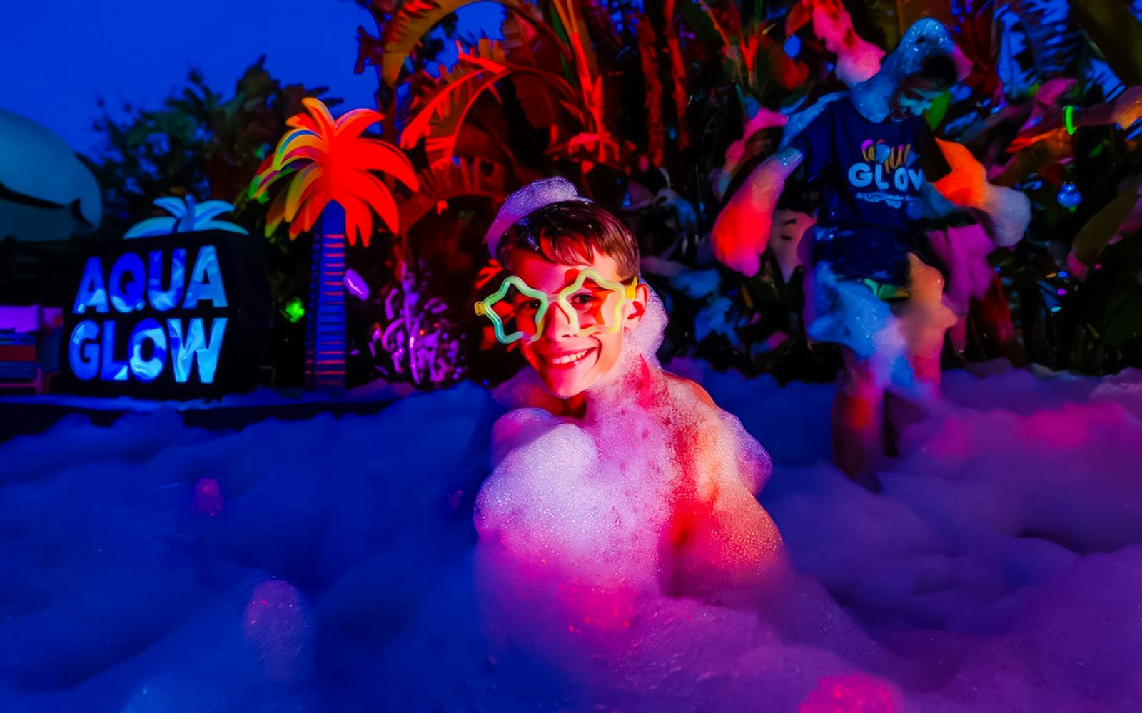 Child enjoying foam party at AquaGlow, Adventure Island Tampa, with colorful lights and palm decor.