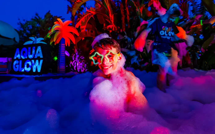 Child enjoying foam party at AquaGlow, Adventure Island Tampa, with colorful lights and palm decor.