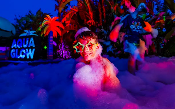 Child enjoying foam party at AquaGlow, Adventure Island Tampa, with colorful lights and palm decor.