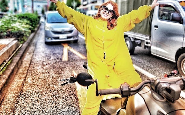 Woman in yellow onesie on go-kart with arms outstretched on Tokyo street.
