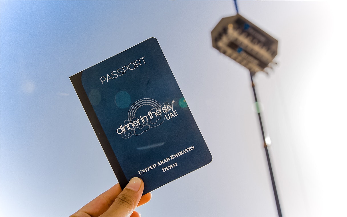 Hand holding a Dinner in the Sky UAE passport with a suspended dining platform in the background.