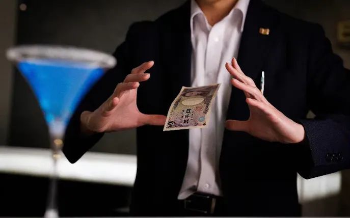 Magician levitating a banknote beside a blue cocktail at BAR21 Tokyo.
