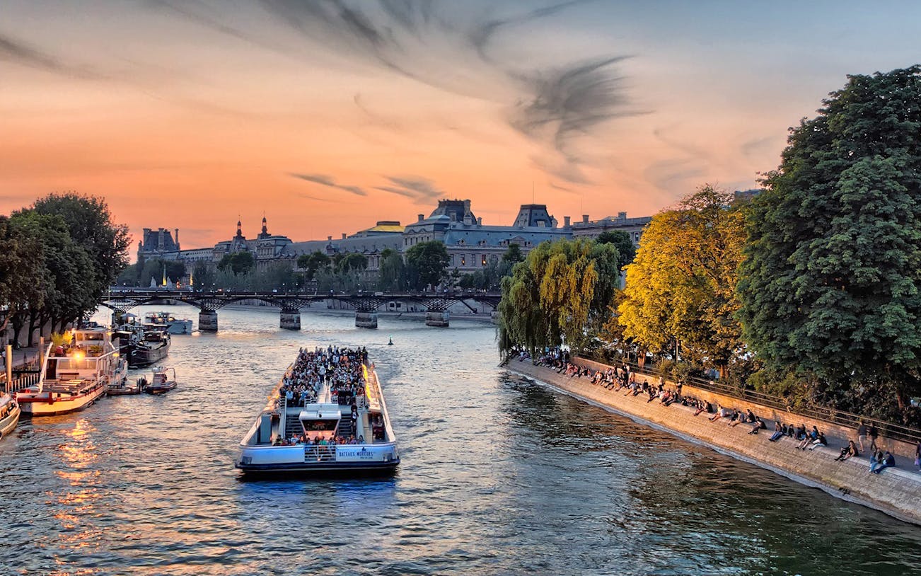 Seine River cruise boat with passengers at sunset in Paris, near historic buildings and trees.