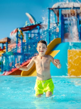 Child playing in a water park pool with colorful slides in the background.