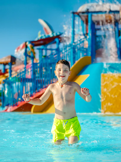 Child playing in a water park pool with colorful slides in the background.