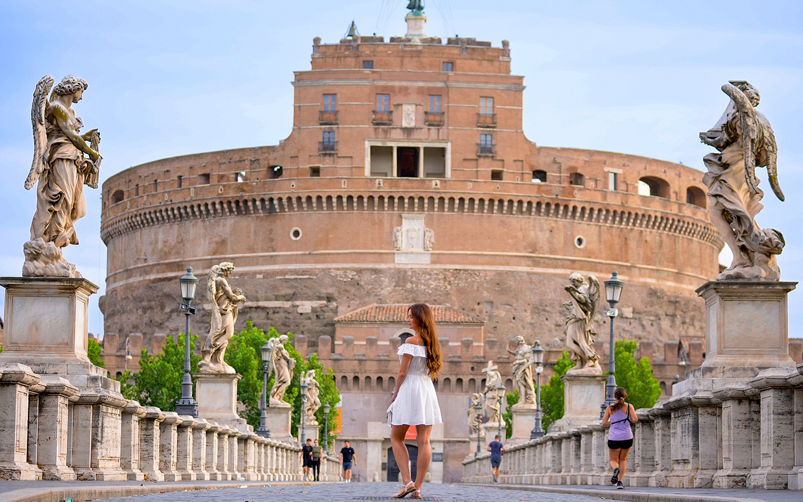 A stylish woman in a white dress strolls across Ponte Sant Angelo, framed by angel statues and the magnificent Castel Sant Angelo in Rome.