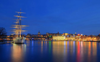 Sailing ship docked in Stockholm harbor at night with city lights reflecting on water.