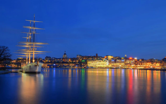 Sailing ship docked in Stockholm harbor at night with city lights reflecting on water.