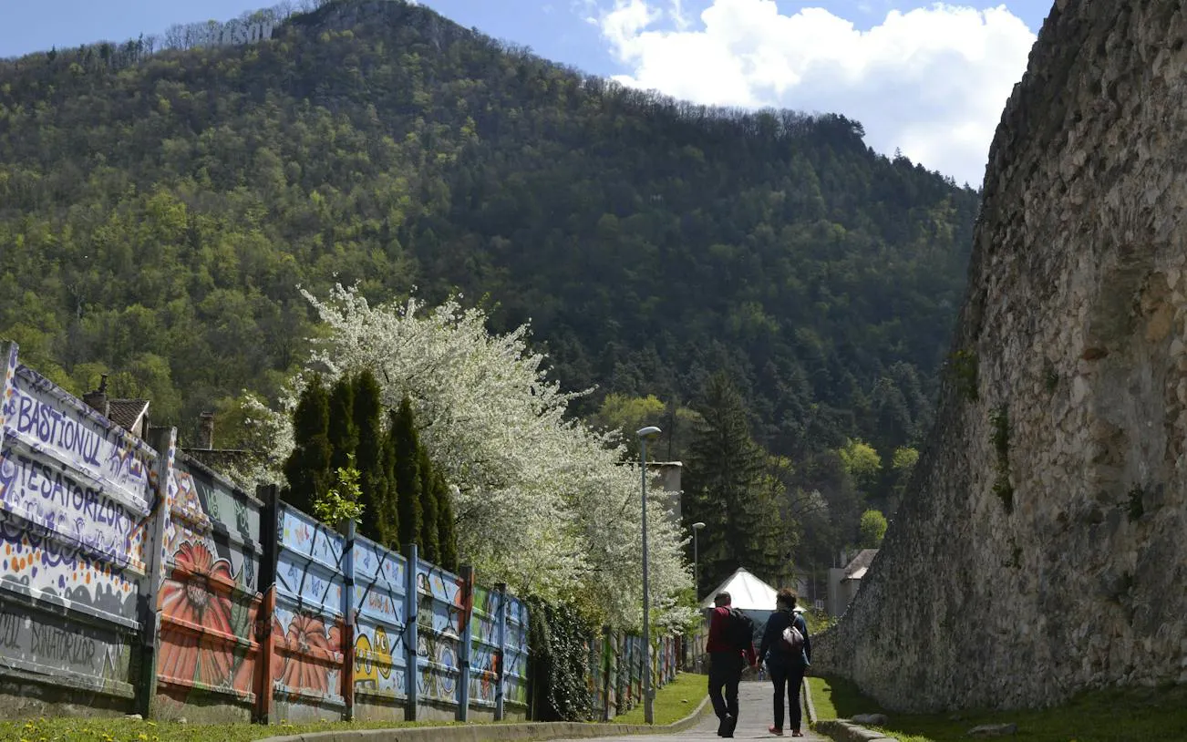 Couple walking along a mural-covered fence and stone wall in Brasov, with a forested hill in the background.