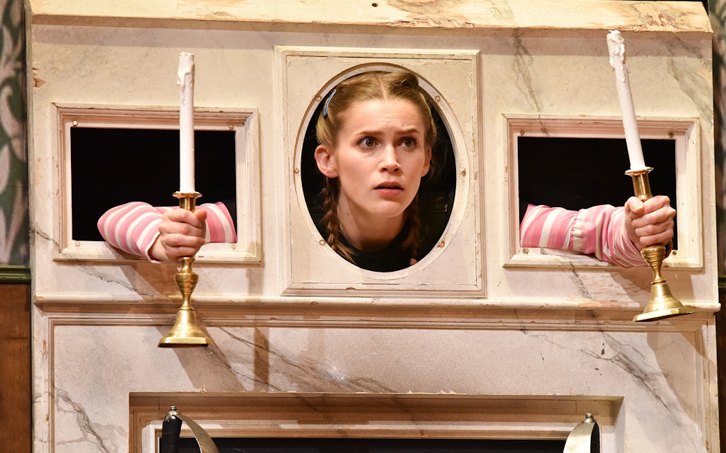 Actor peering through a wall with candlesticks in "The Play That Goes Wrong" scene.