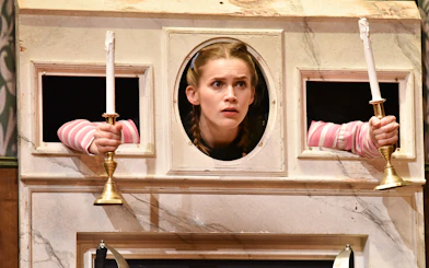Actor peering through a wall with candlesticks in "The Play That Goes Wrong" scene.